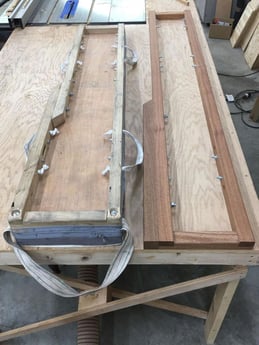Photo of two wooden piano moving boards on a workbench in a workshop. The boards are equipped with handles and are used for securing and transporting pianos. Various woodworking tools and materials are visible in the background.