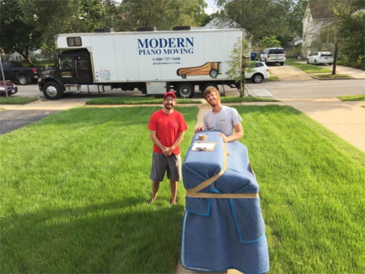 Two men stand on a lawn with a piano wrapped in a blue blanket. A Modern Piano Moving truck is in the background