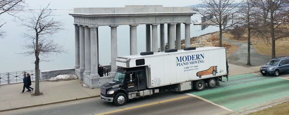 Modern Piano Moving truck parked near a historic columned structure by the water.
