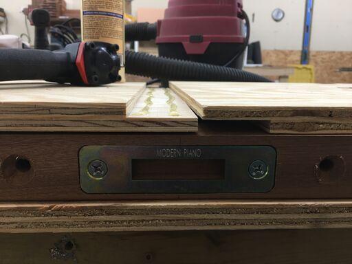 Close-up photo of a wooden workbench in a workshop, showing a metal plate labeled 'MODERN PIANO' attached to the wood. Various tools and equipment, including a vacuum and glue, are visible in the background.
