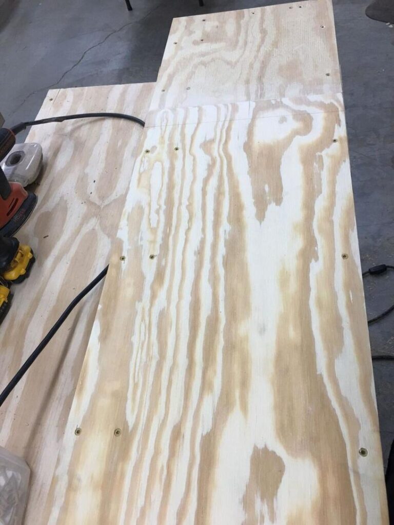 Photo of a piece of plywood on a workbench with several screws partially drilled in. An electric sander and other tools are visible on the left side of the workbench."