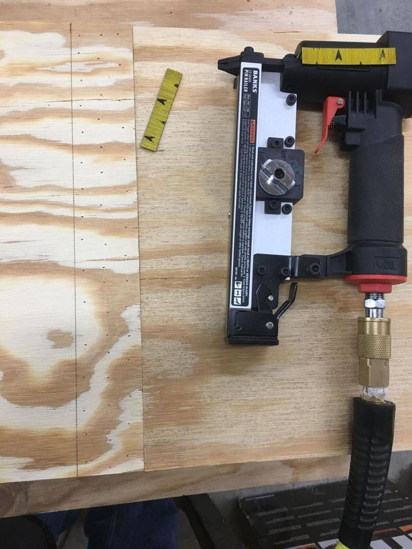 Close-up photo of a pneumatic nail gun on a wooden workbench, alongside a piece of plywood marked with a measuring tape. The setup is prepared for woodworking in a workshop.
