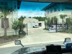 Photo taken from inside a truck cab, showing the view of a storage facility with palm trees, gated entry, and buildings with garage doors. Signs and a directional arrow are visible outside the facility.