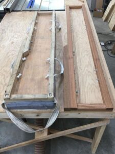 Photo of two wooden piano moving boards on a workbench in a workshop. The boards are equipped with handles and are used for securing and transporting pianos. Various woodworking tools and materials are visible in the background.
