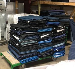 Photo of a large stack of moving blankets folded neatly on a green dolly in a warehouse. Shelving and other moving supplies are visible in the background.