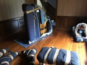 Photo of an upright piano wrapped in moving blankets and secured with straps in a room with hardwood floors. Several padded and wrapped items are on the floor, and a man is kneeling, wrapping another item in the background.
