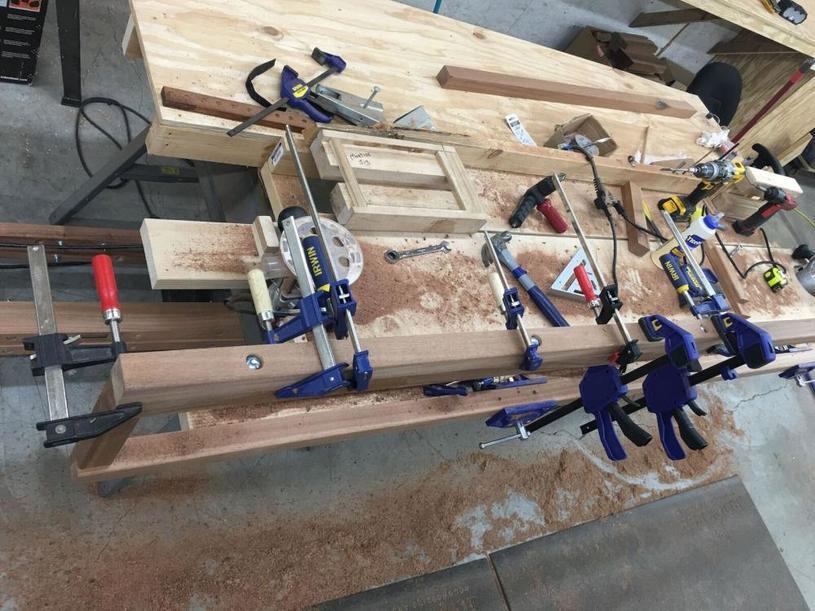 Photo of a workbench in a woodworking workshop with various tools and clamps securing wood pieces. The bench is covered with sawdust and features tools such as hammers, drills, and measuring instruments.