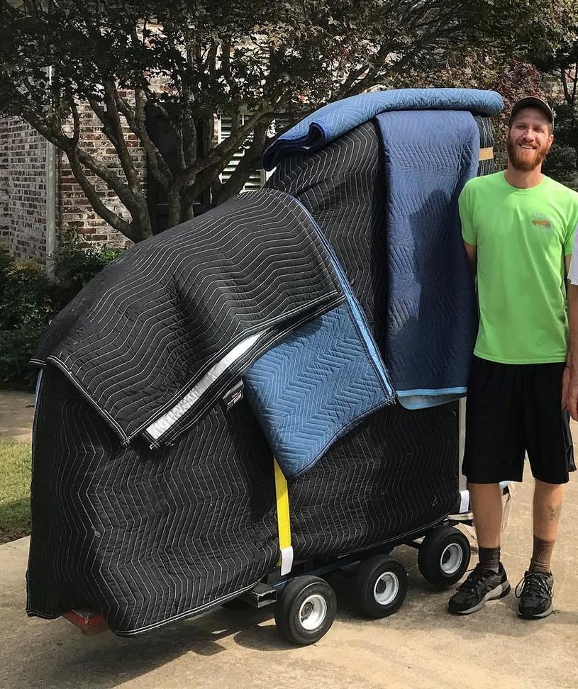 Photo of a man standing next to an upright piano wrapped in protective moving blankets and secured with straps on a piano dolly. The man is smiling and dressed in a green shirt and black shorts, with trees and a brick building in the background.