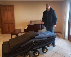 Photo of a man standing next to a dolly loaded with folded moving blankets inside a room with a piano and a wooden door in the background. The man is dressed in a jacket and cap, smiling at the camera.