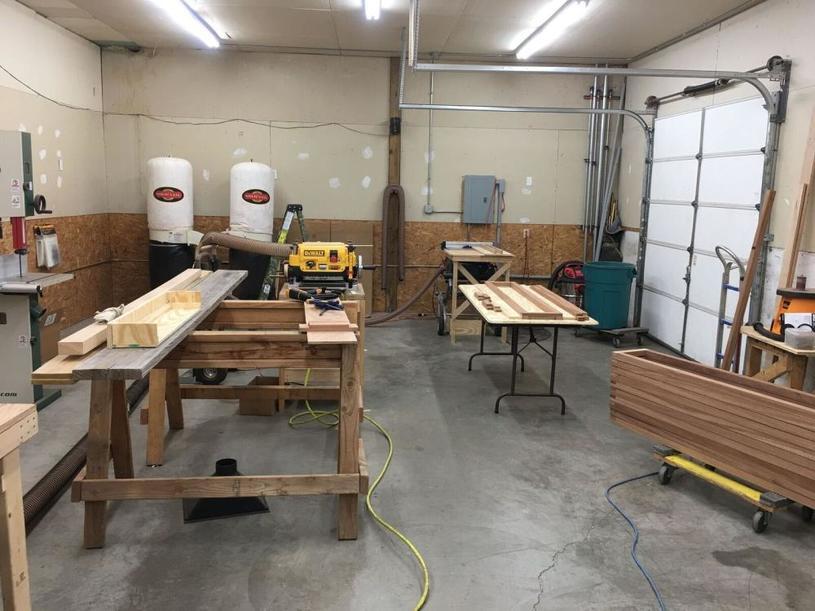 "Photo of a woodworking workshop with various tools and equipment, including a table saw, workbenches, and wood pieces in progress. The space has a concrete floor, high ceilings, and a partially open garage door.