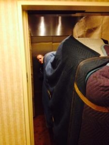 Photo of a person standing inside an elevator, partially obscured by large, padded furniture pieces that are being moved. The furniture is covered with protective moving blankets.
