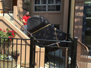 Photo of a man moving a large, padded piano up an outdoor staircase. The piano is covered with protective blankets and secured with straps. The scene is set in front of a building entrance with a black metal fence and flower pots.