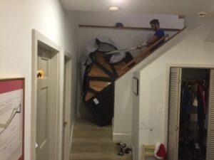 Photo of a man moving a large piano up a narrow indoor staircase with a tight turn. The piano is partially wrapped in protective blankets, and the man is guiding it with straps. The scene is set in a hallway with a closet and a framed picture on the wall