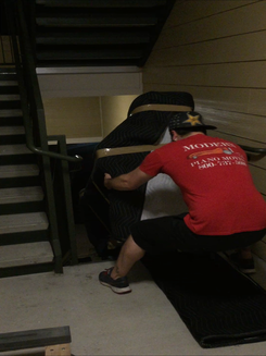 Photo of a man moving a large, padded piano up a narrow stairwell in an apartment building. The man is wearing a red shirt and is guiding the piano with straps. The stairwell has green handrails and beige walls.