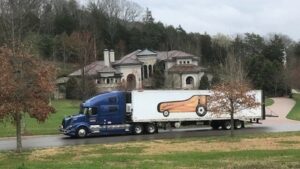 Photo of a large moving truck with a grand piano graphic on the side, parked in front of a luxurious mansion surrounded by trees and a winding driveway.
