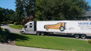 Photo of a large moving truck with a grand piano graphic on the side, driving on a suburban street with well-manicured lawns and trees.