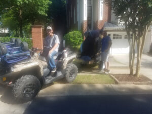 Photo of two men moving a piano using an ATV in front of a suburban house with a garage. One man is seated on the ATV, while the other is guiding the piano on a small trailer.