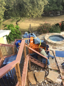 Photo of three people moving a large, padded piano down a set of outdoor deck stairs. The piano is wrapped in protective blankets and secured with straps. The scene is set in a backyard with a hot tub, stone patio, and surrounding greenery.