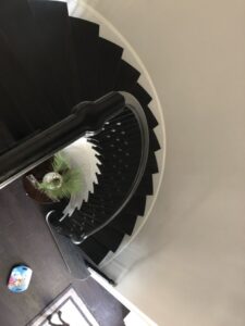 Photo taken from the top of a curved staircase looking down. The dark wooden steps and black handrail create a striking spiral pattern. At the bottom of the stairs, a small table with a plant and a children's toy on the floor are visible.