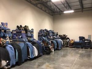 Photo of a warehouse interior with numerous items wrapped in moving blankets and stacked against the walls. The space is large with high ceilings and concrete floors.