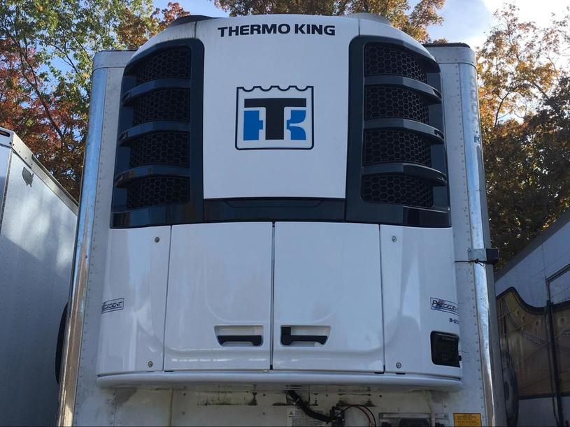 Photo of the front of a Thermo King refrigeration unit mounted on a trailer, with the Thermo King logo prominently displayed. Trees and other trailers are visible in the background.