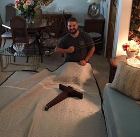 Photo of a man sitting on the floor, wrapping a large item with a moving blanket in a well-furnished living room. The room features a dining table, chairs, and floral arrangements.