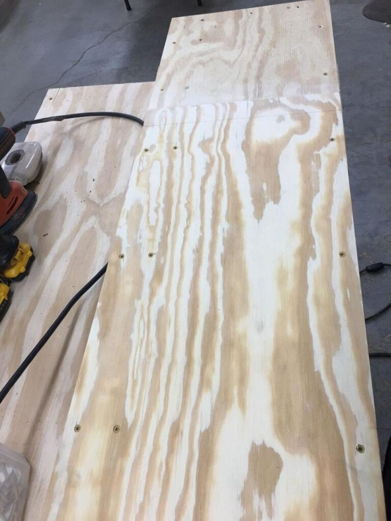 Photo of a piece of plywood on a workbench with several screws partially drilled in. An electric sander and other tools are visible on the left side of the workbench."