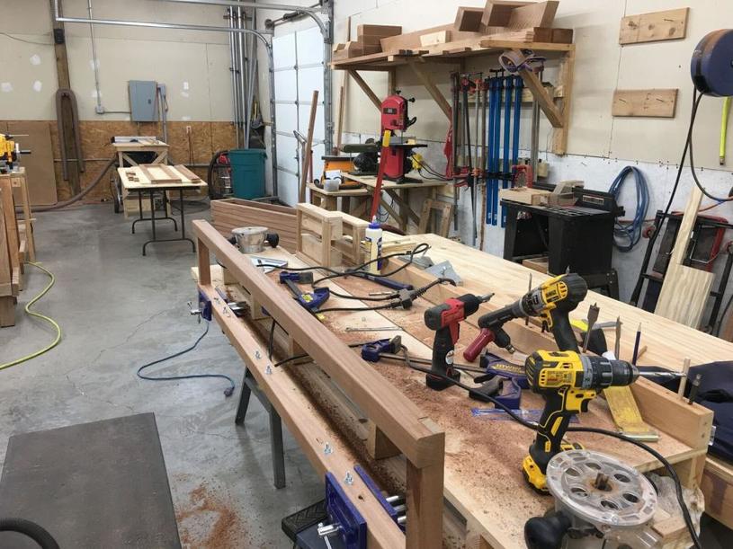 Photo of a busy woodworking workshop with various tools and equipment on workbenches, including drills, clamps, and wood pieces. Shelves with additional tools and materials are visible in the background.