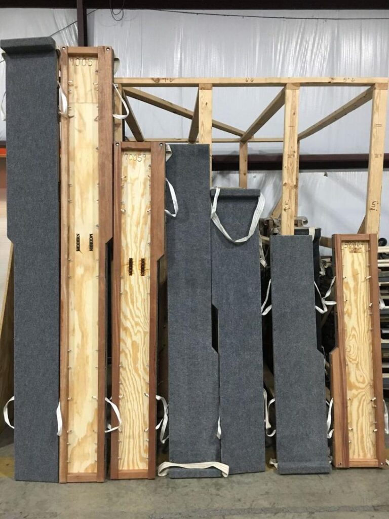 Photo of multiple wooden piano moving boards standing upright in a workshop. The boards are covered with gray fabric on one side and have fabric handles for carrying.
