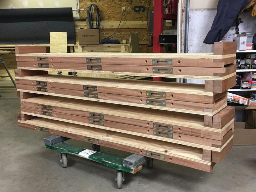 Photo of several stacked wooden piano moving boards with metal plates, labeled 'MODERN PIANO,' arranged on a green dolly in a workshop. The background shows various woodworking tools and materials