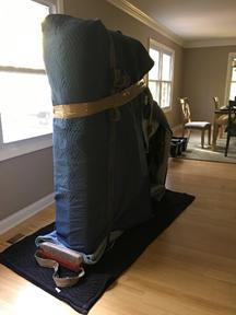 Photo of an upright piano wrapped in protective moving blankets and secured with straps, placed on a dolly inside a room with hardwood floors and large windows.