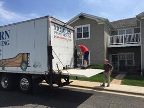 Photo of two men unloading a piano from a Modern Piano Moving truck using a liftgate. The scene is set in front of a residential building with balconies and well-maintained landscaping.